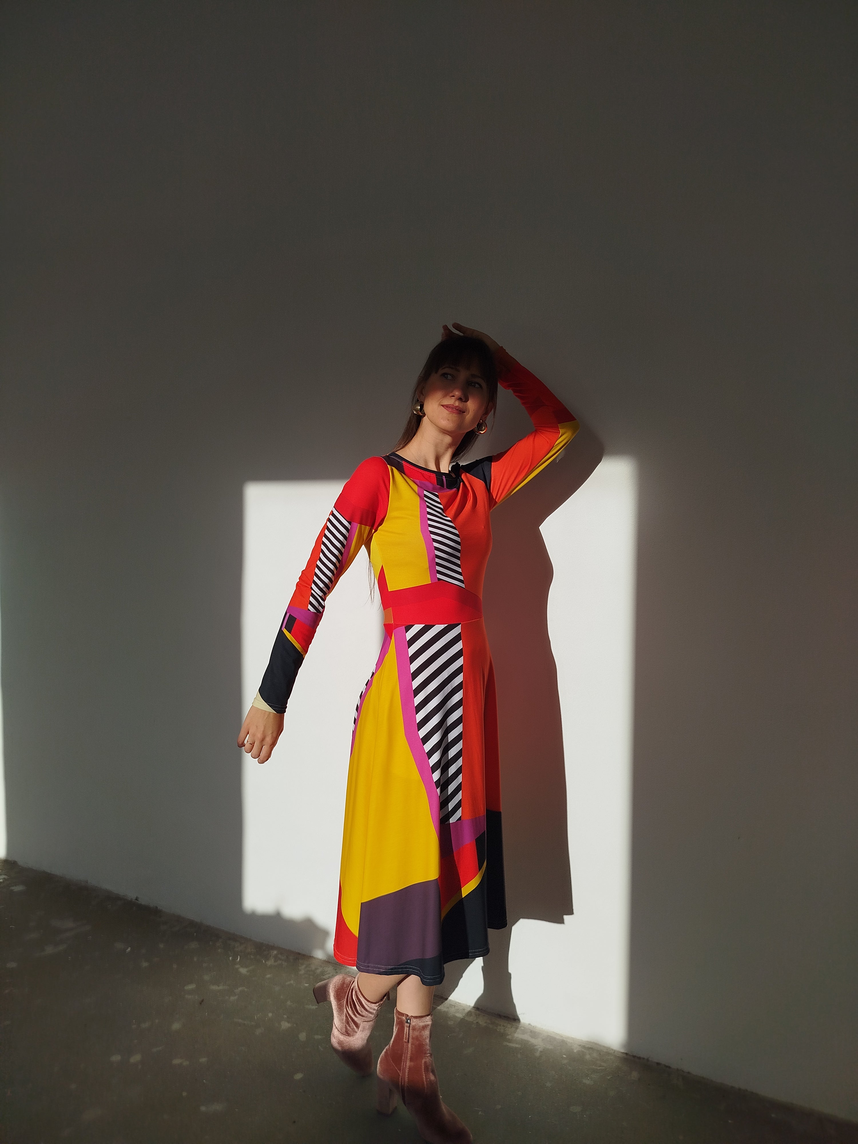 Person wearing a colorful orange dress with bold black and white stripes, artistic statement fashion design, standing against a white wall with dramatic shadows, modern wearable art style, creative contemporary clothing.