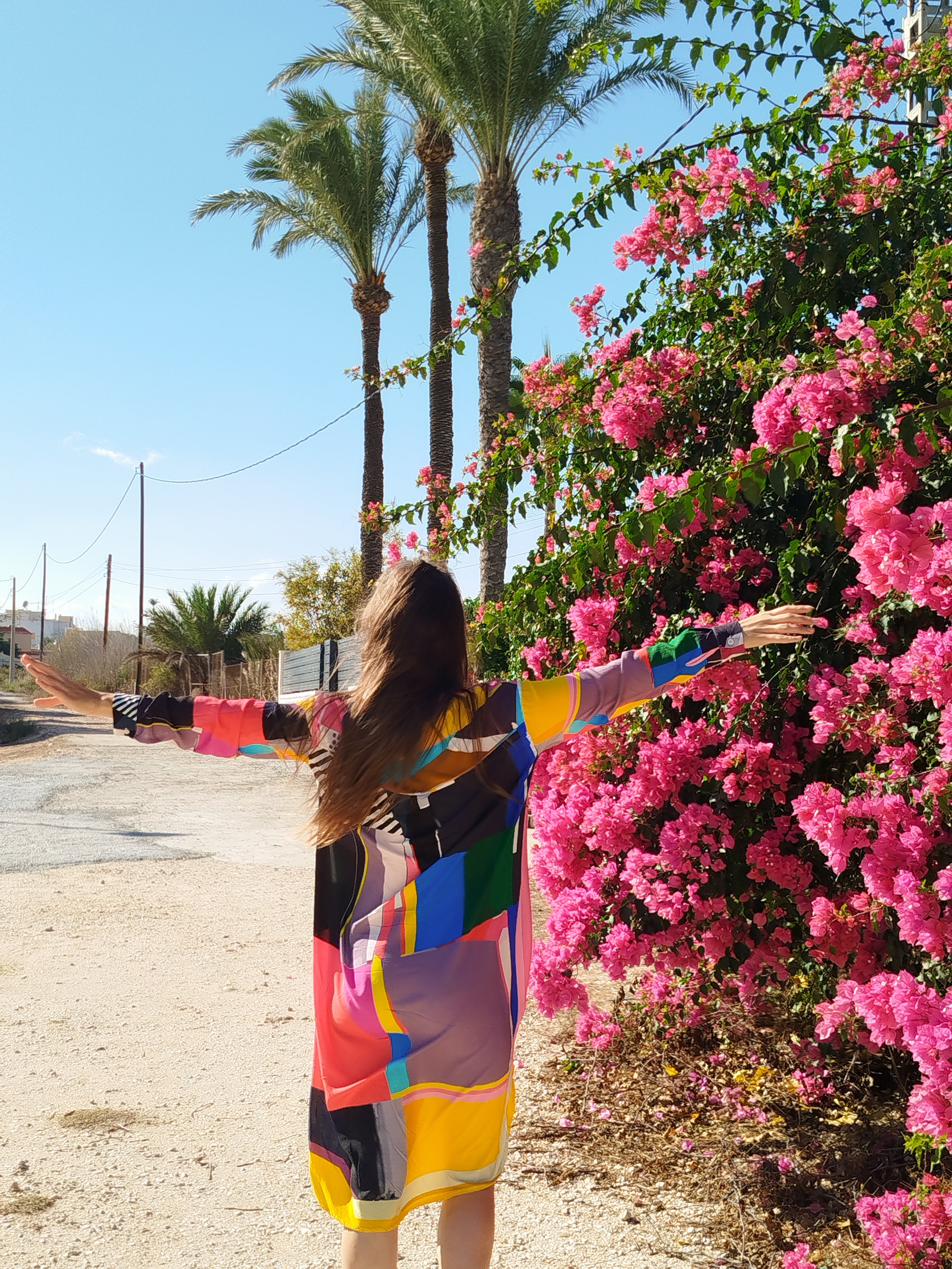 Model wearing a colorful artistic dress, standing next to a flowering bush with vibrant blossoms, palm trees in the background, tropical outdoor setting, modern wearable art fashion, bold creative style inspired by nature