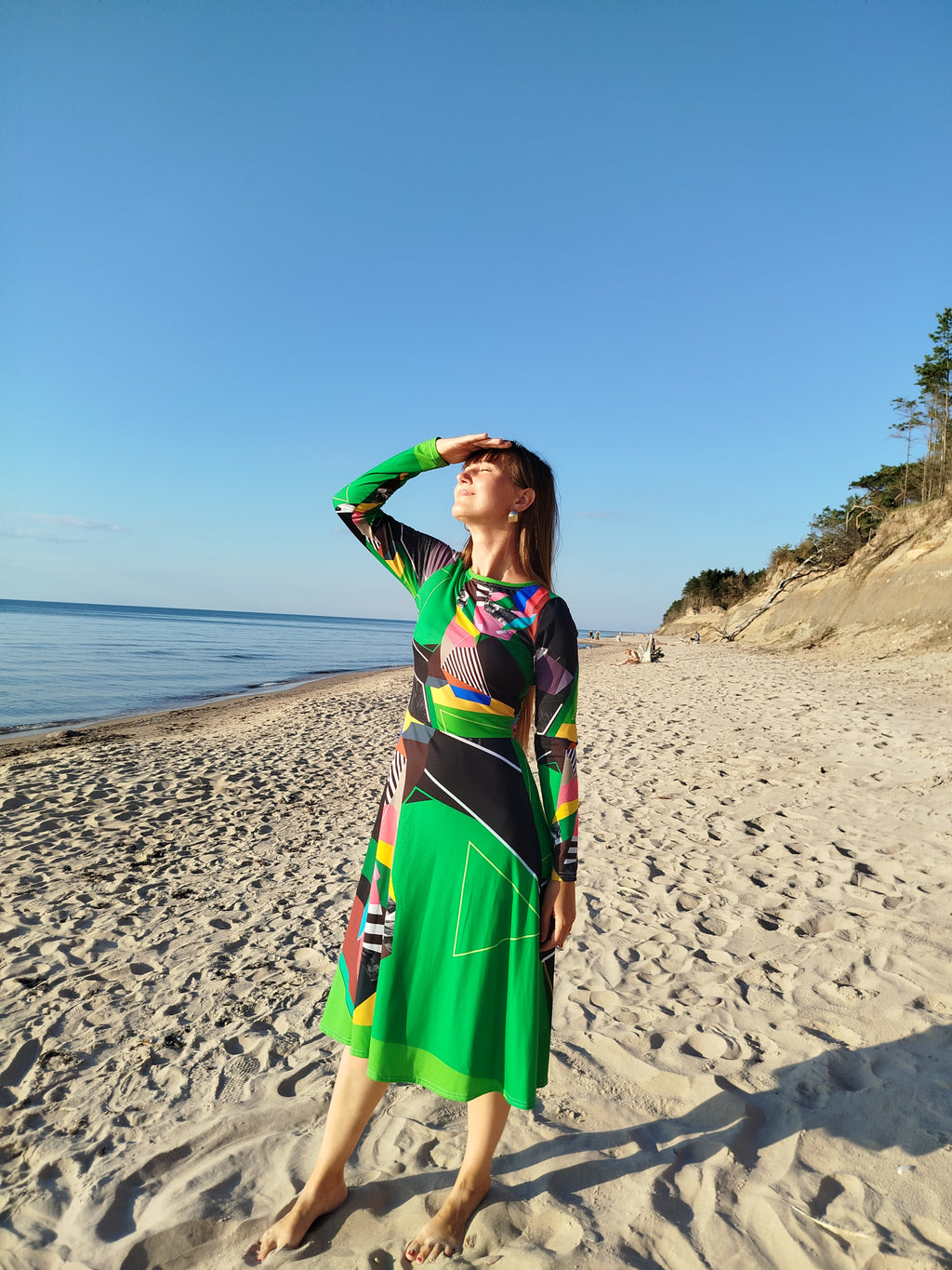 Woman wearing a green abstract midi dress with long sleeves, bold artistic abstract pattern, modern statement fashion design, photographed on a sandy beach with a clear blue sky, contemporary wearable art dress for women