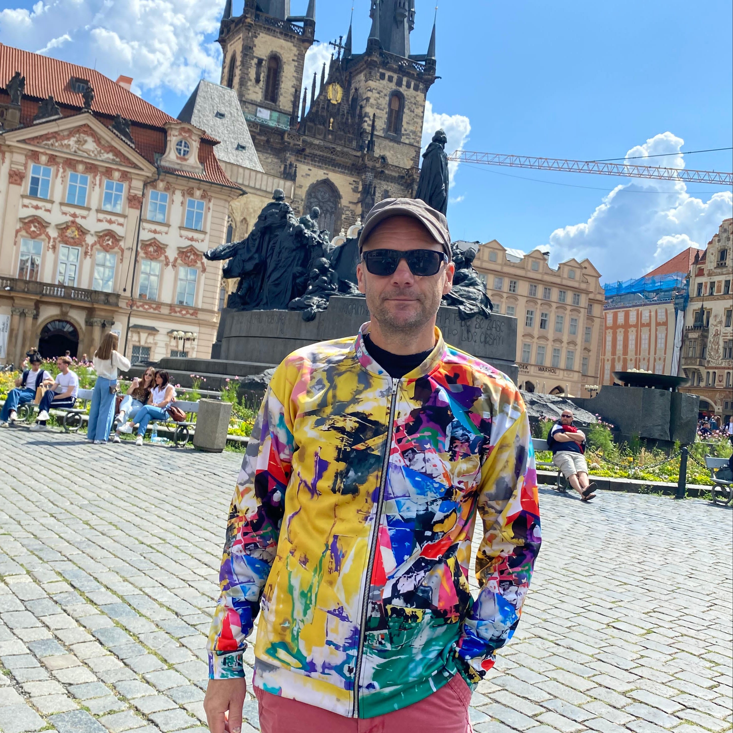 Man in a colorful jacket and pink pants standing in a Prague square with historic architecture.