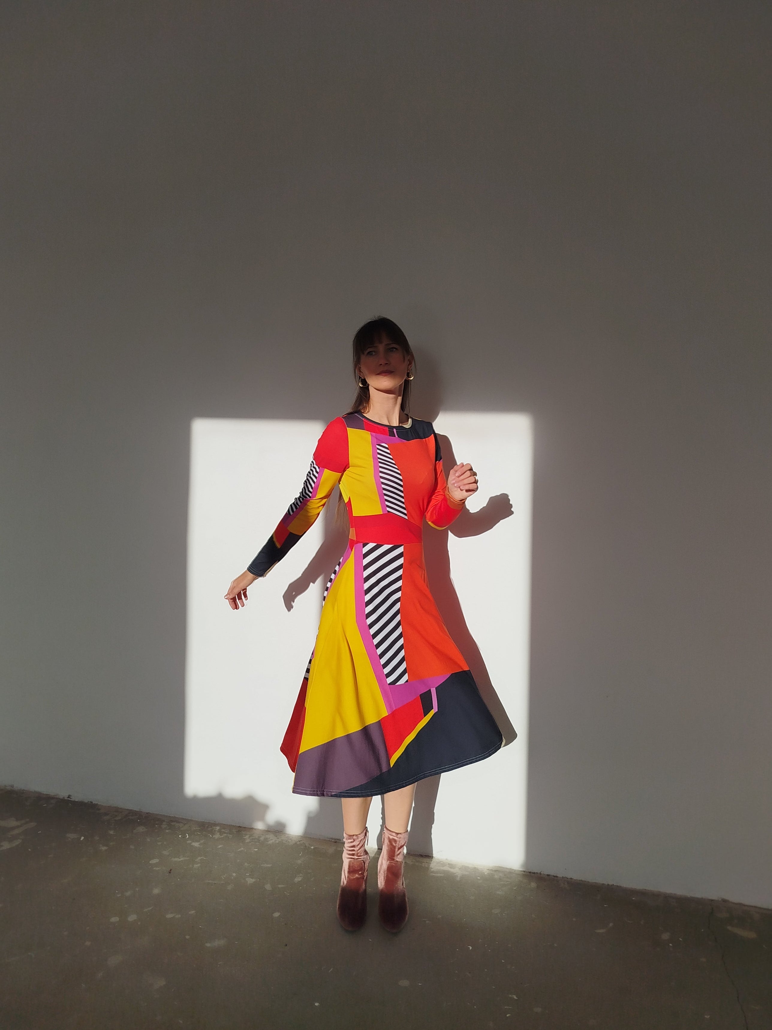 Model stepping lightly in a contemporary color-block dress blending crimson, sunflower yellow, and monochrome stripes, the silhouette animated by motion.