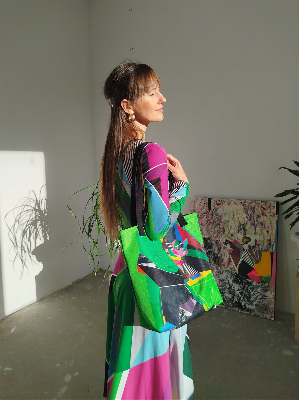 Woman in a green abstract midi dress holding a colorful tote bag in a room with plants and artwork.