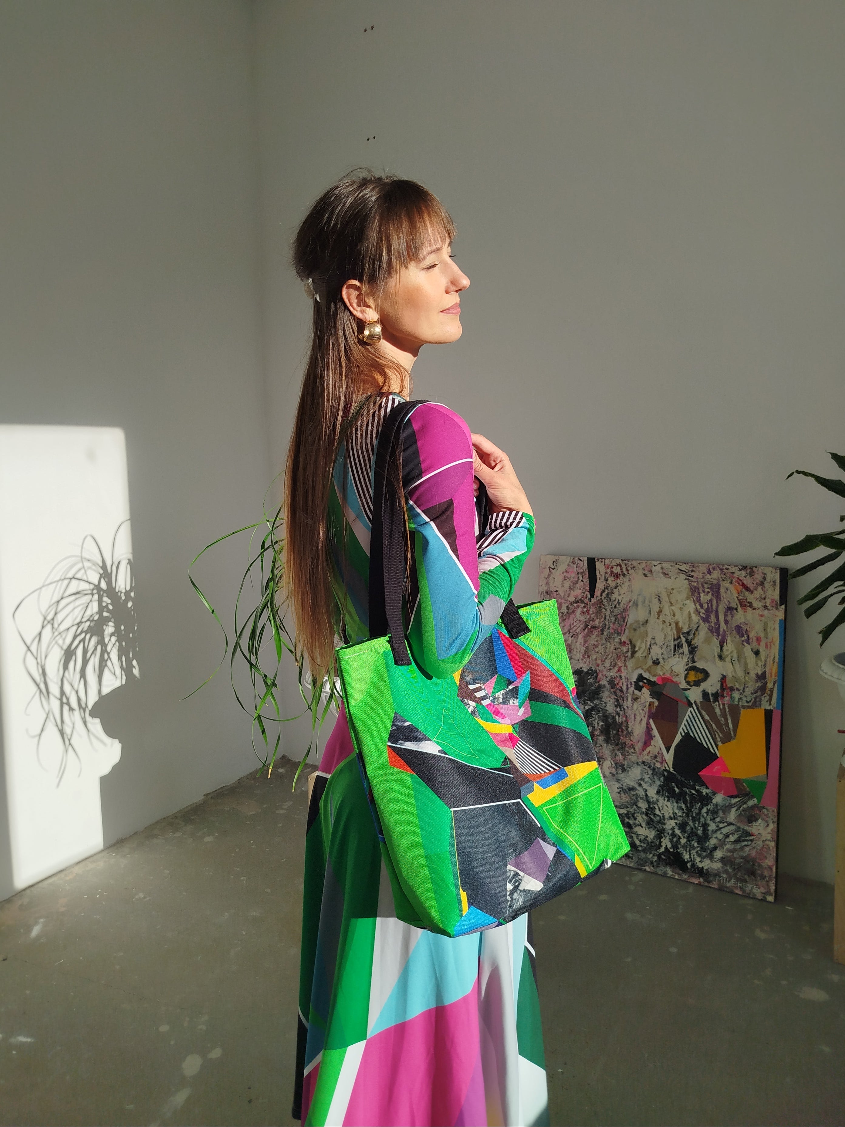 Woman in a green abstract midi dress holding a colorful tote bag in a room with plants and artwork.