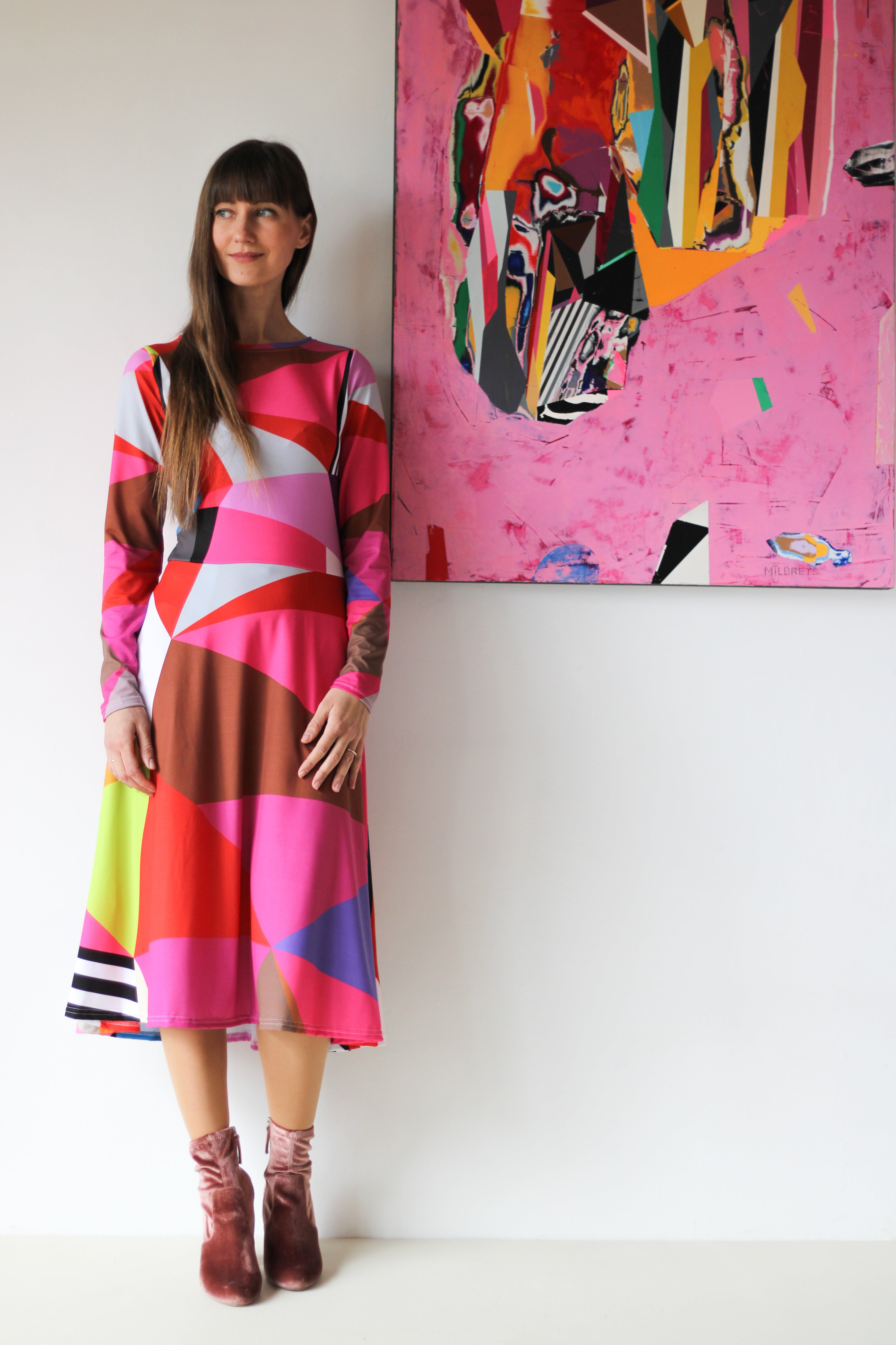 Model wearing a colorful pink geometric dress with bold abstract patterns, standing next to a vibrant abstract painting, artistic fashion inspired by modern art, expressive wearable art dress, creative contemporary style photographed in an art gallery setting