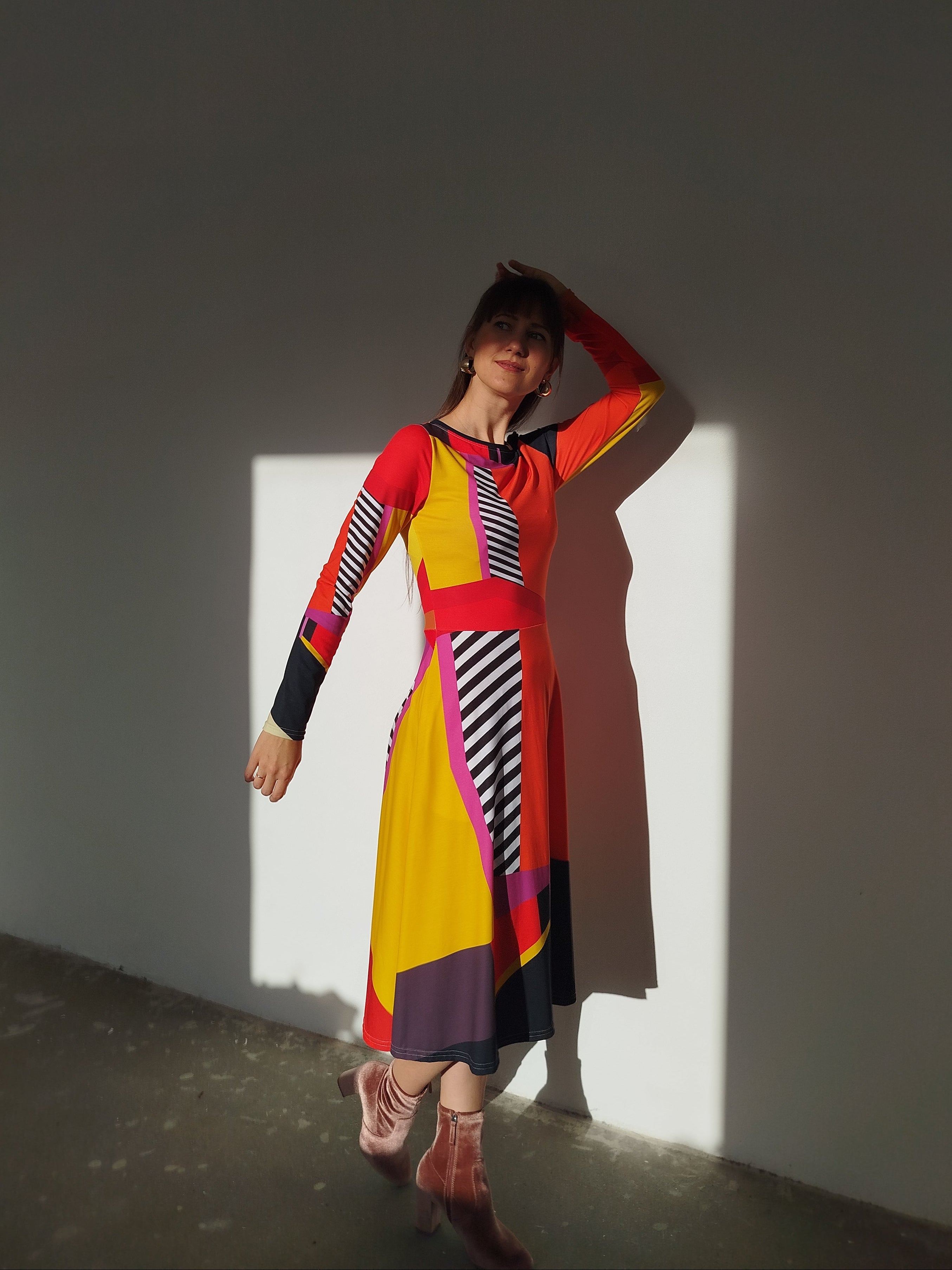 Bold long-sleeve midi dress in vibrant red, yellow, black, and pink color blocks with graphic stripes, fitted waist, and flowing skirt—modern art-inspired statement piece lit by dramatic sunlight.