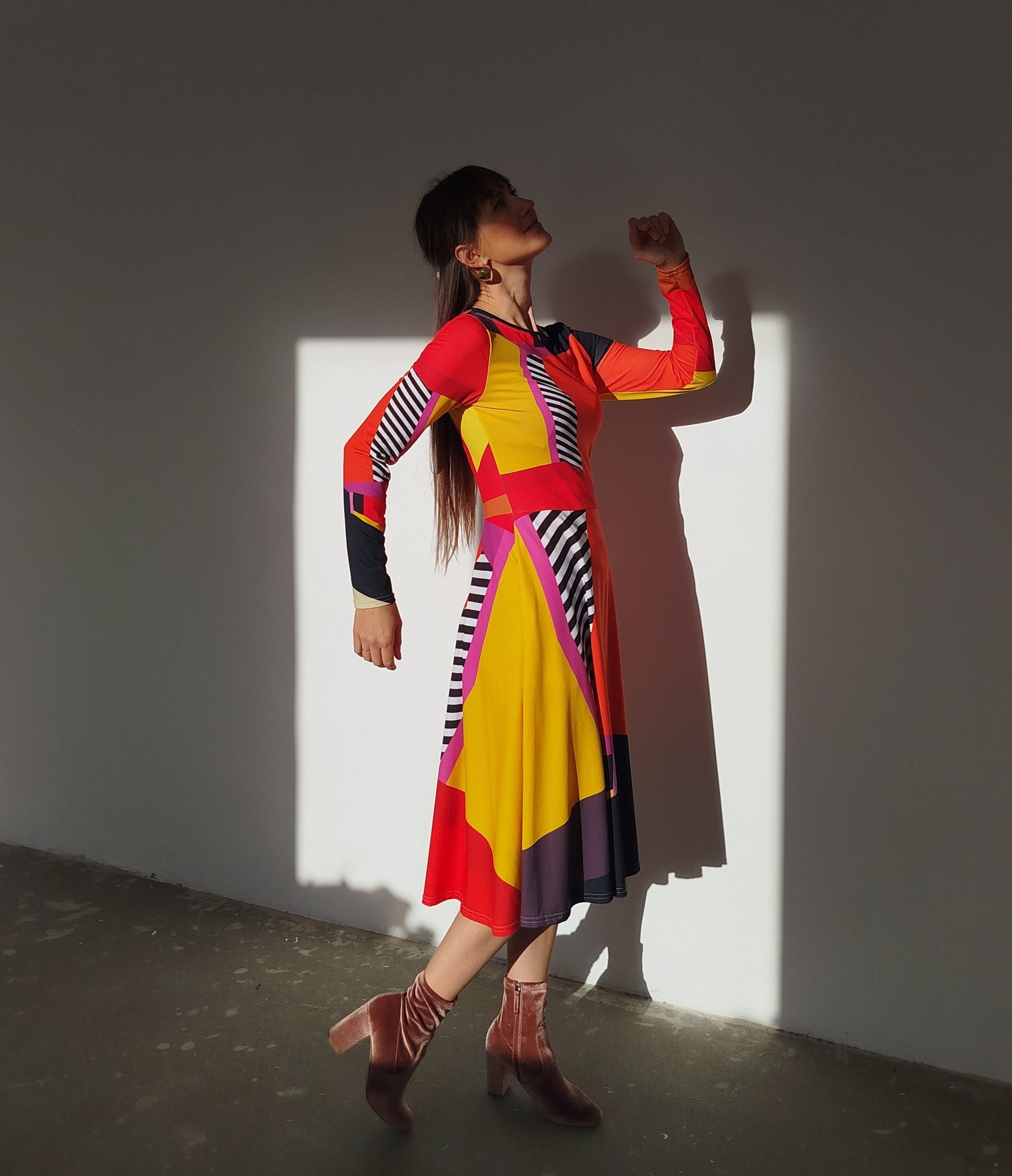 Model dancing in a bold long-sleeve midi dress with vibrant red, yellow, black, and pink color blocking and graphic stripes, the flowing skirt catching sunlight as she moves.