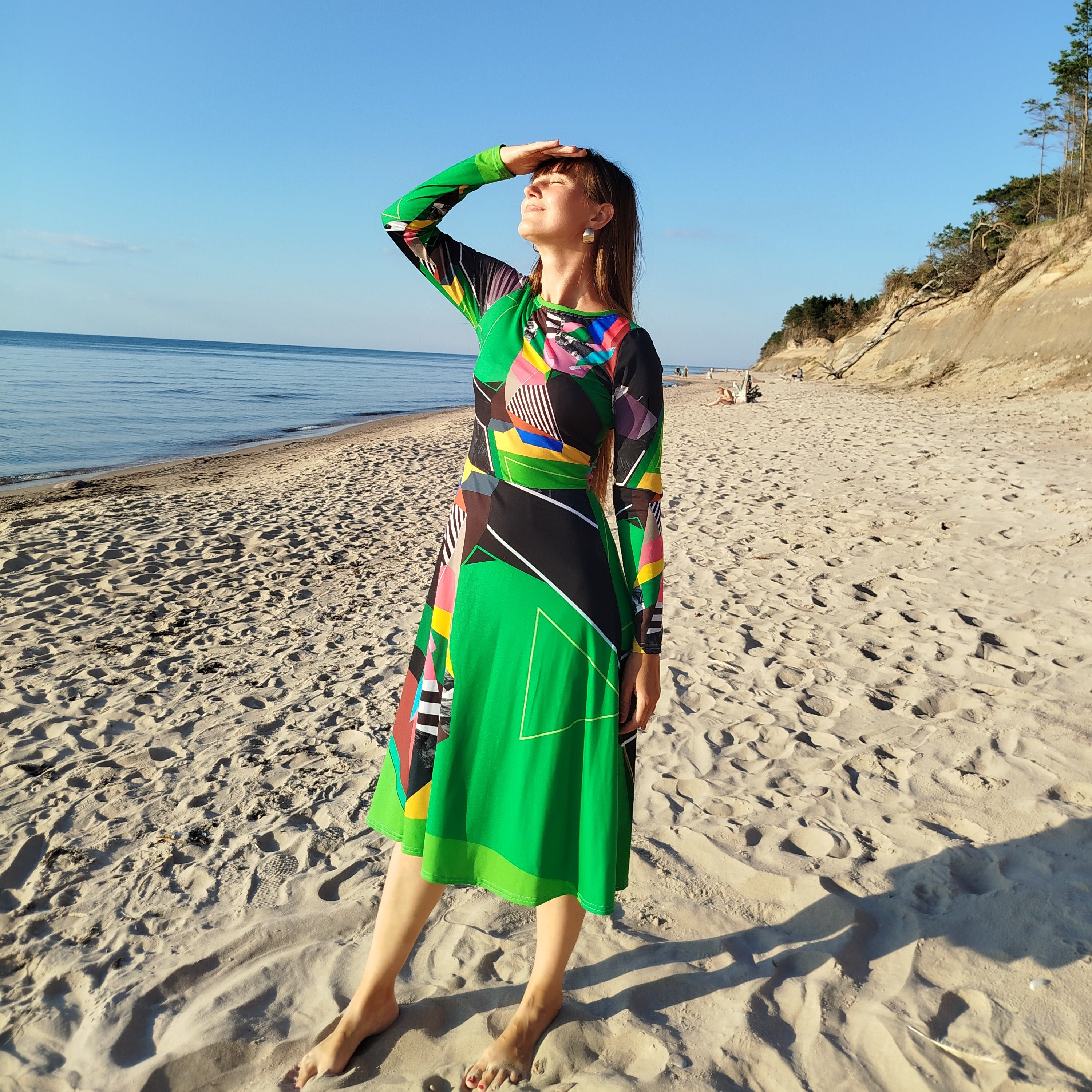 Woman wearing a green abstract midi dress with long sleeves, bold artistic abstract pattern, modern statement fashion design, photographed on a sandy beach with a clear blue sky, contemporary wearable art dress for women