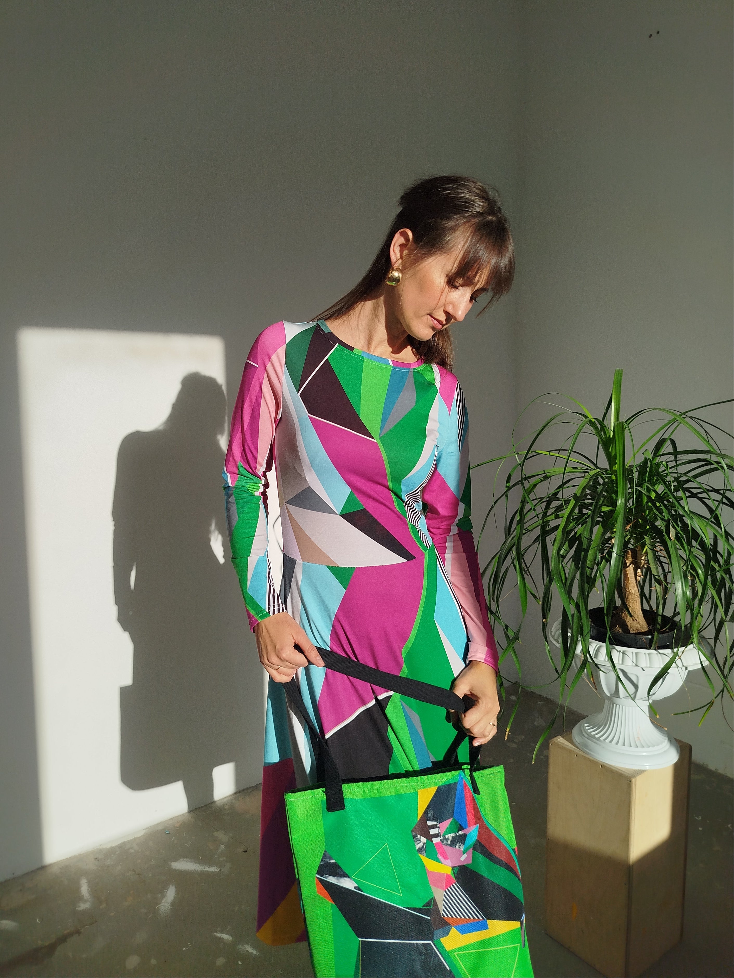 Woman in a colorful green dress holding a green bag in a room with a plant and light fixture.